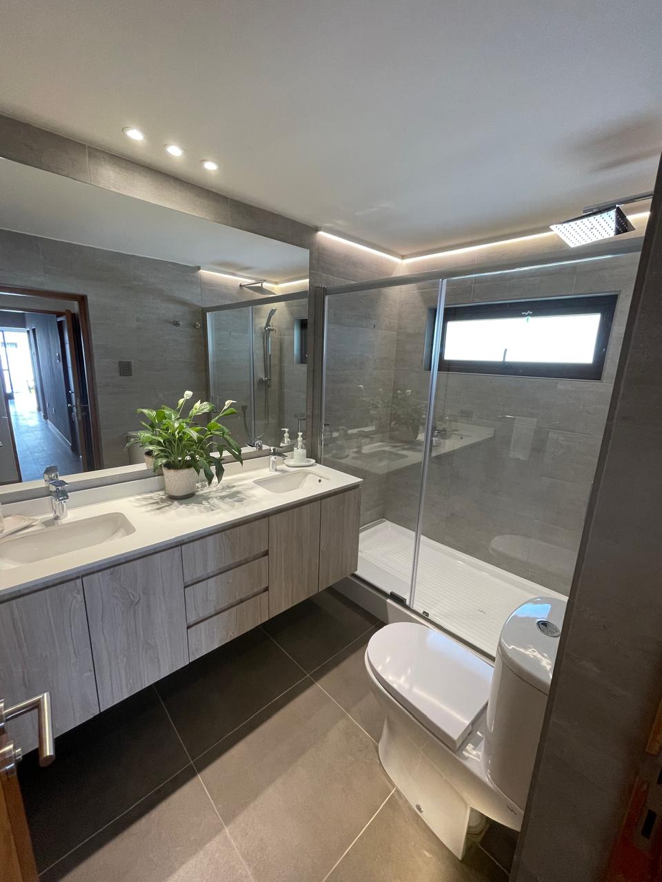 Baño principal en tonos piedra con doble vanitorio, ducha con lluvia y luz indirecta