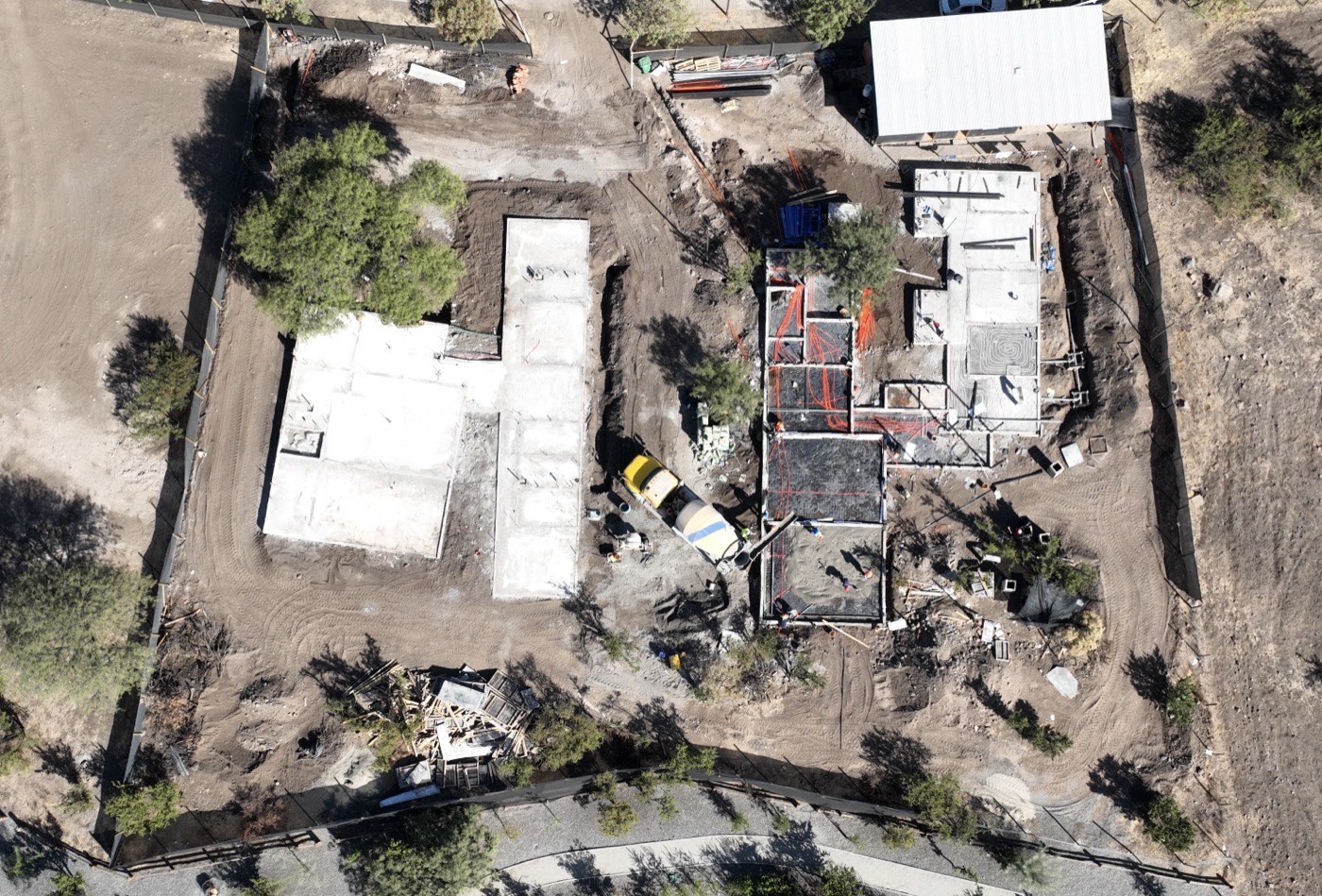 Obra en ejecución vista desde el aire: fundaciones y losas de dos viviendas, camión mixer y cuadrilla trabajando entre árboles